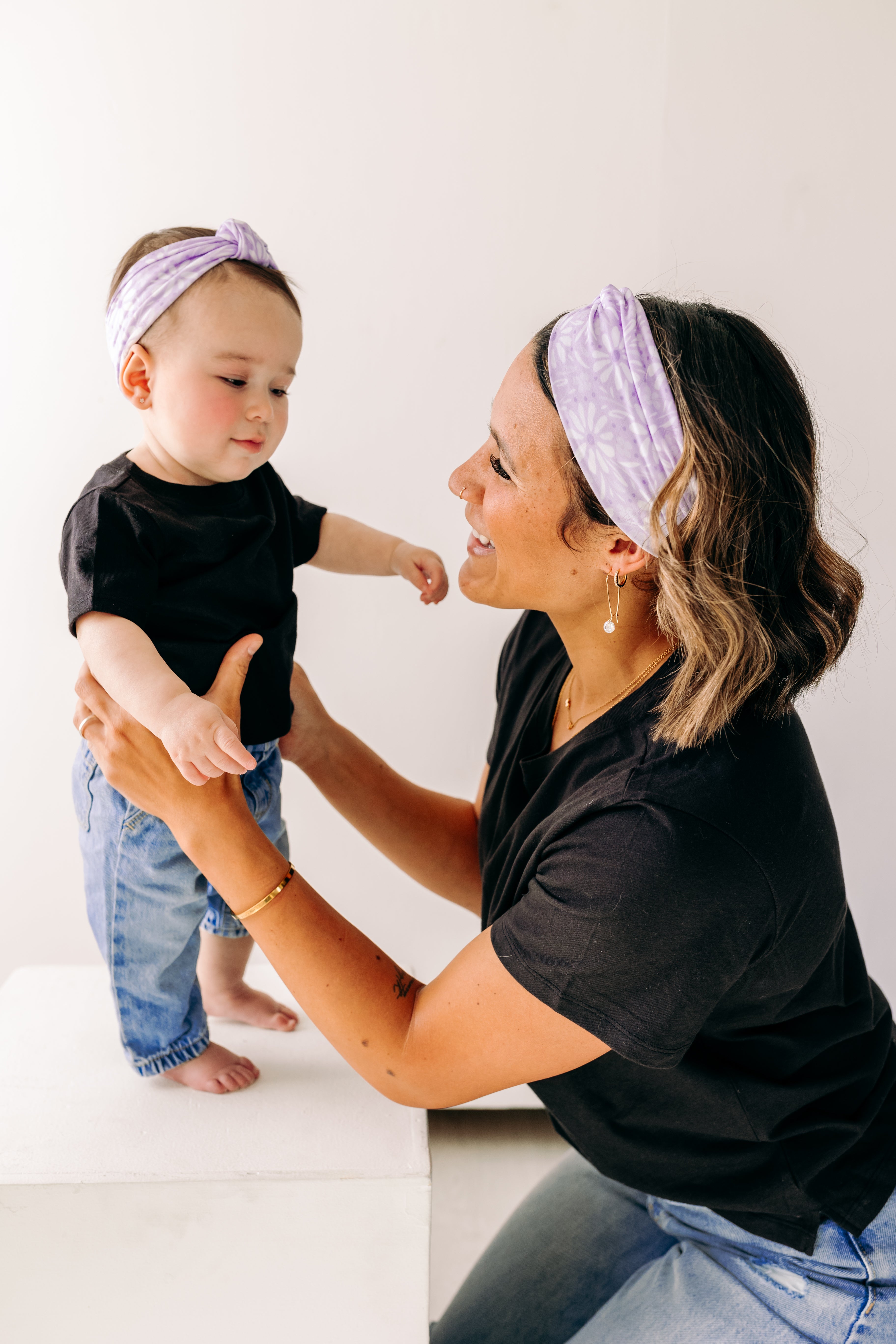 A woman smiles holding a baby, both wearing Darby Floral on Purple headbands. Adjustable bow tie style for all ages, machine washable, ultra-soft poly-knit blend. Designed in Springfield, MO.