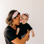 A woman smiles holding a baby, both wearing Riley Floral on Ivory headbands. Adjustable bow tie style for all ages, machine washable, ultra-soft poly-knit blend. Designed in Springfield, MO.