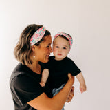 A woman smiles holding a baby, both wearing Riley Floral on Ivory headbands. Adjustable bow tie style for all ages, machine washable, ultra-soft poly-knit blend. Designed in Springfield, MO.