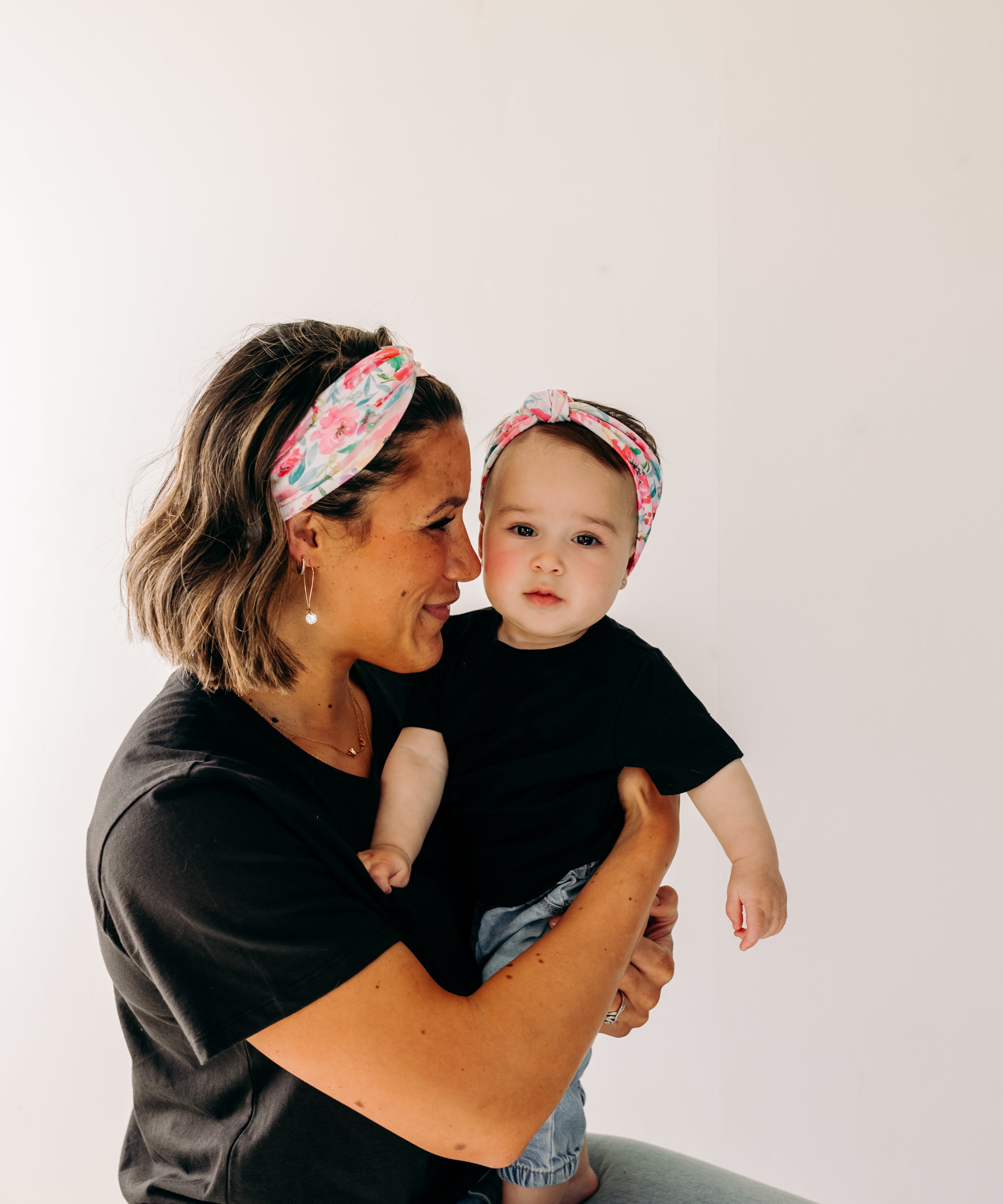A woman smiles holding a baby, both wearing Riley Floral on Ivory headbands. Adjustable bow tie style for all ages, machine washable, ultra-soft poly-knit blend. Designed in Springfield, MO.