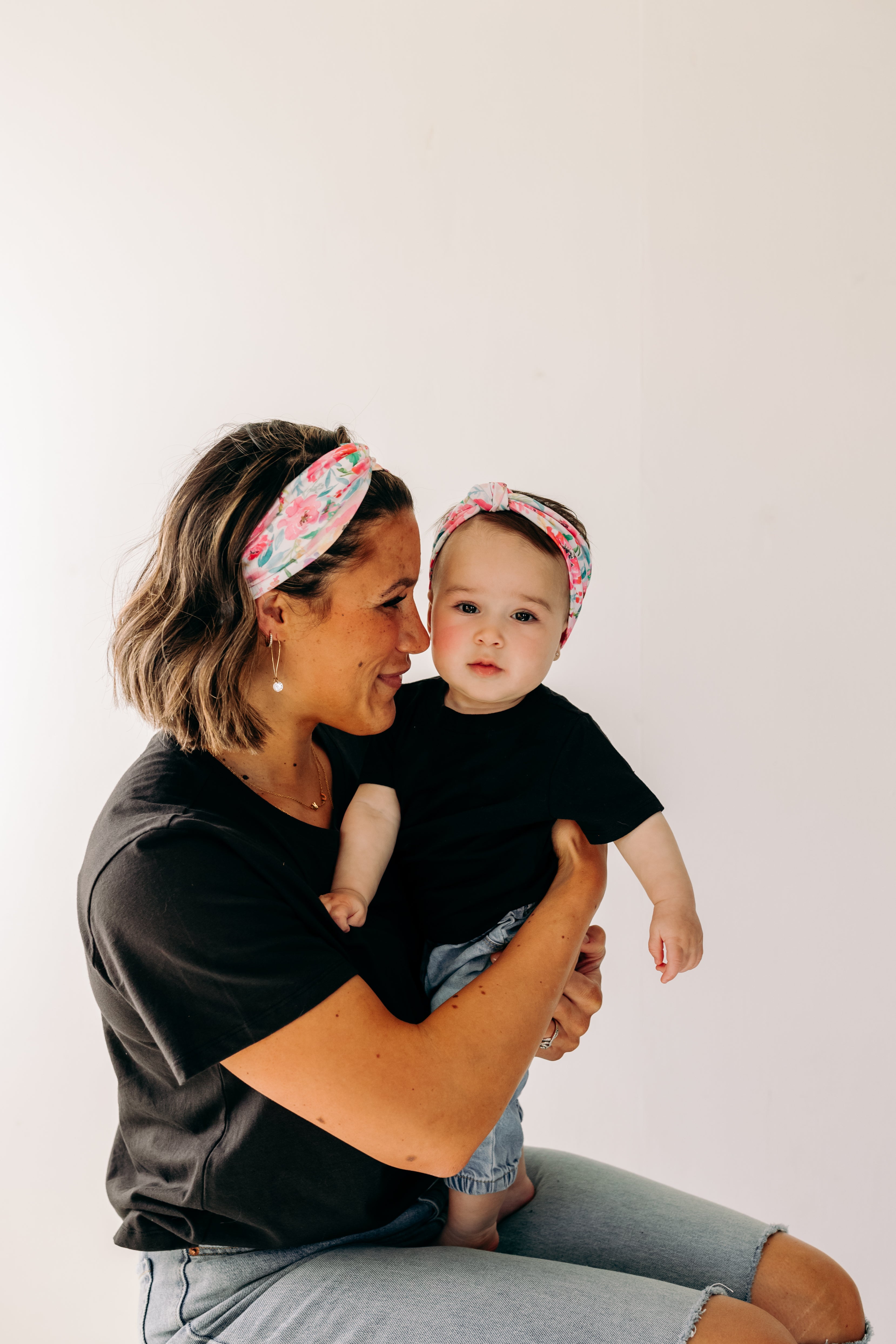 A woman smiles holding a baby, both wearing Riley Floral on Ivory headbands. Adjustable bow tie style for all ages, machine washable, ultra-soft poly-knit blend. Designed in Springfield, MO.
