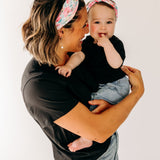 A woman smiles holding a baby, both wearing Riley Floral on Ivory headbands. Adjustable bow tie style for all ages, machine washable, ultra-soft poly-knit blend. Designed in Springfield, MO.