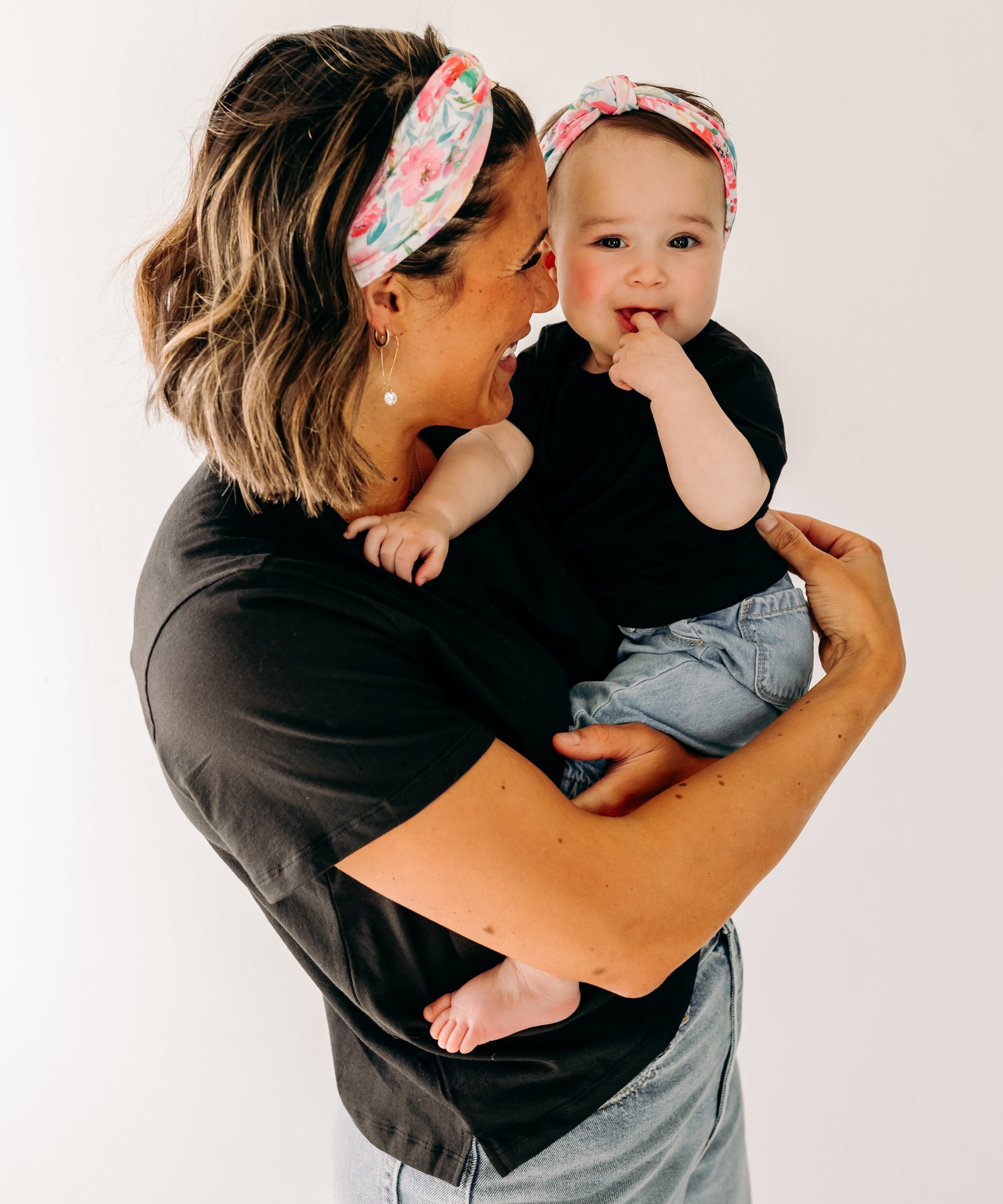 A woman smiles holding a baby, both wearing Riley Floral on Ivory headbands. Adjustable bow tie style for all ages, machine washable, ultra-soft poly-knit blend. Designed in Springfield, MO.