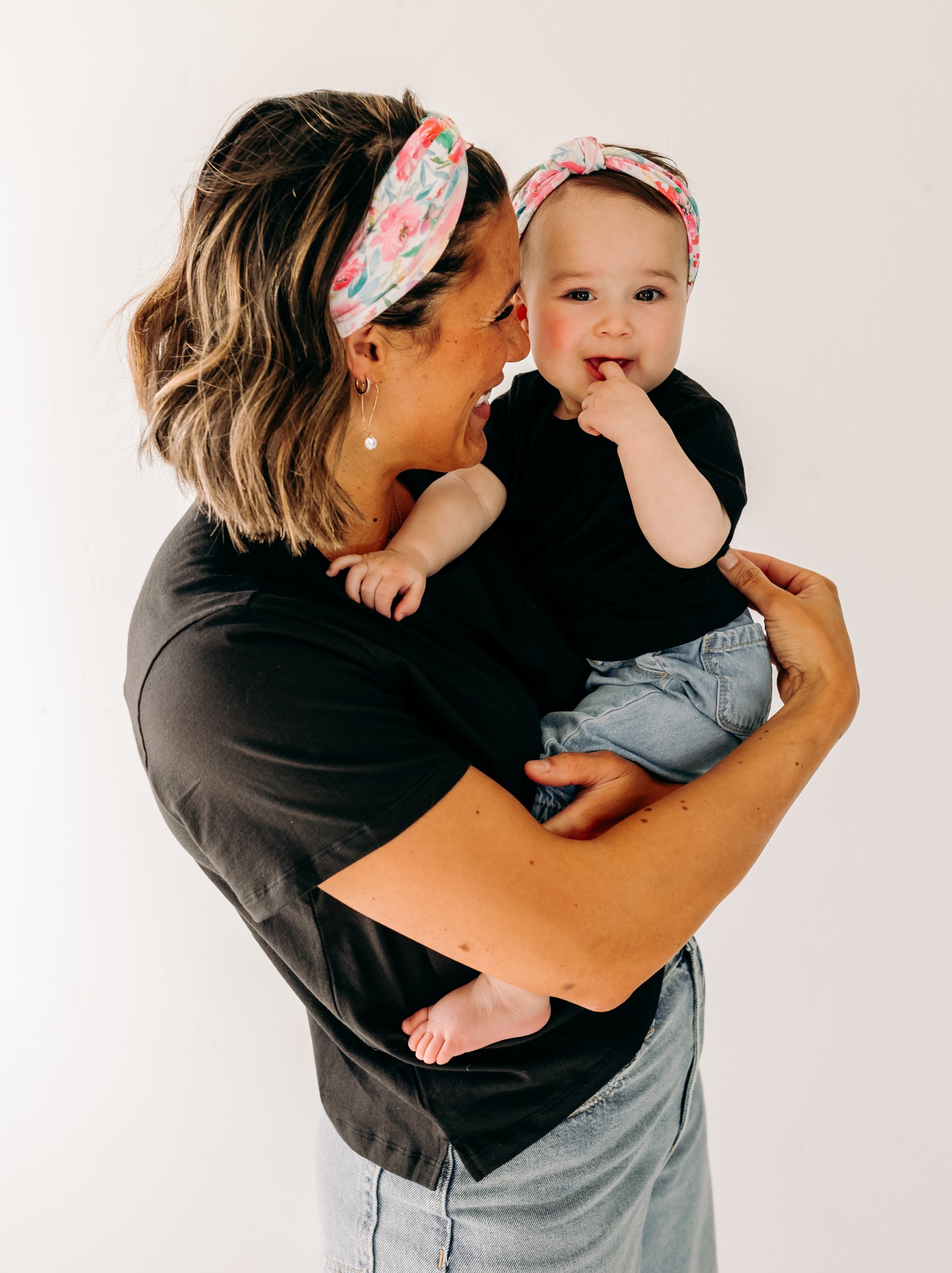 A woman smiles holding a baby, both wearing Riley Floral on Ivory headbands. Adjustable bow tie style for all ages, machine washable, ultra-soft poly-knit blend. Designed in Springfield, MO.