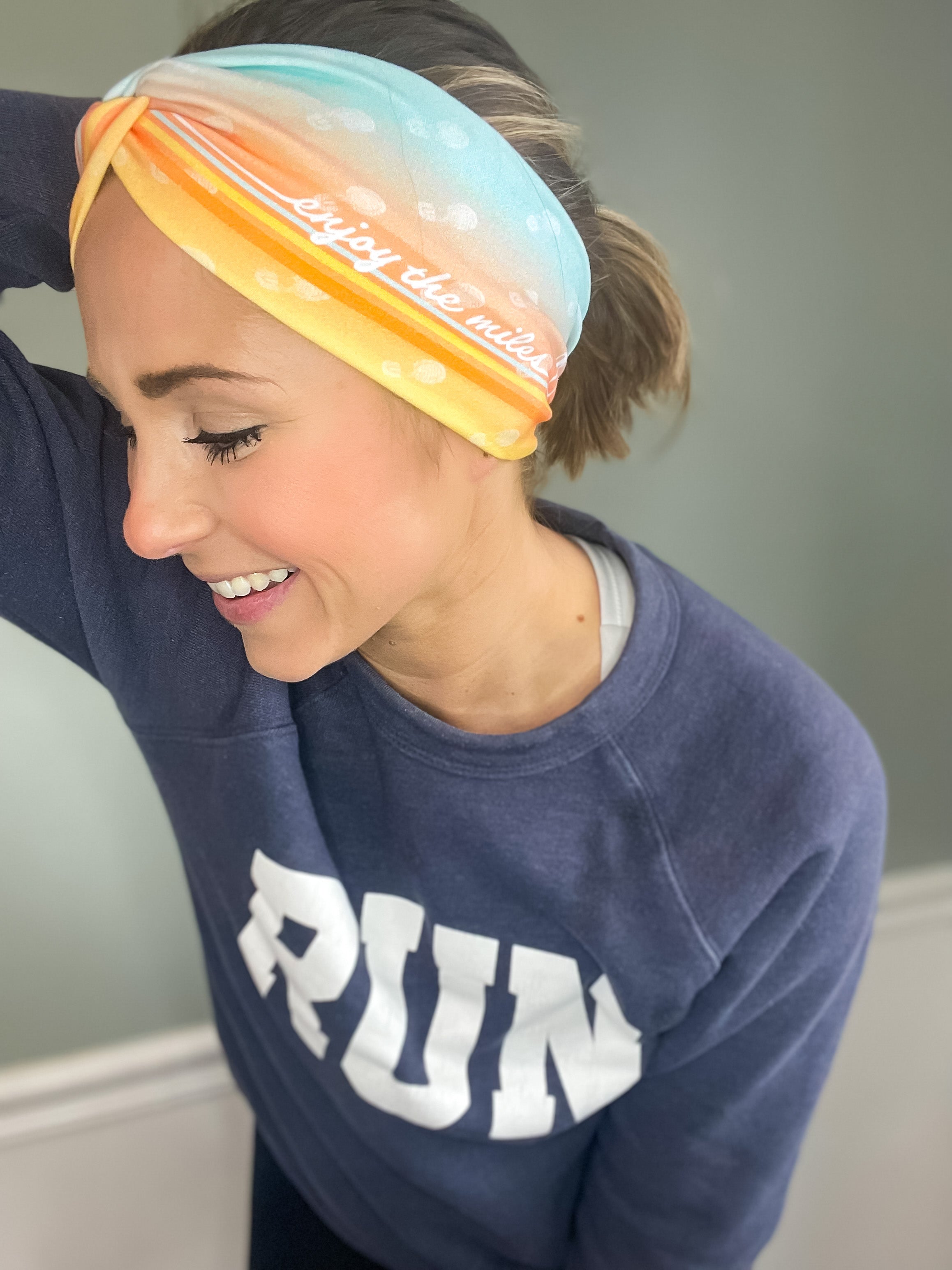 Adult women's gradient headband with yellow, orange and blue with allover white shoe prints, three yellow, orange and white horizontal lines and the white script saying "enjoy the miles"