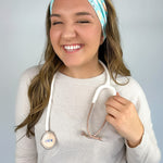 Adult women's blue-teal headband with yellow smiley faces, pink daisies, muted rainbows, and pink stethoscopes with a heart on the end