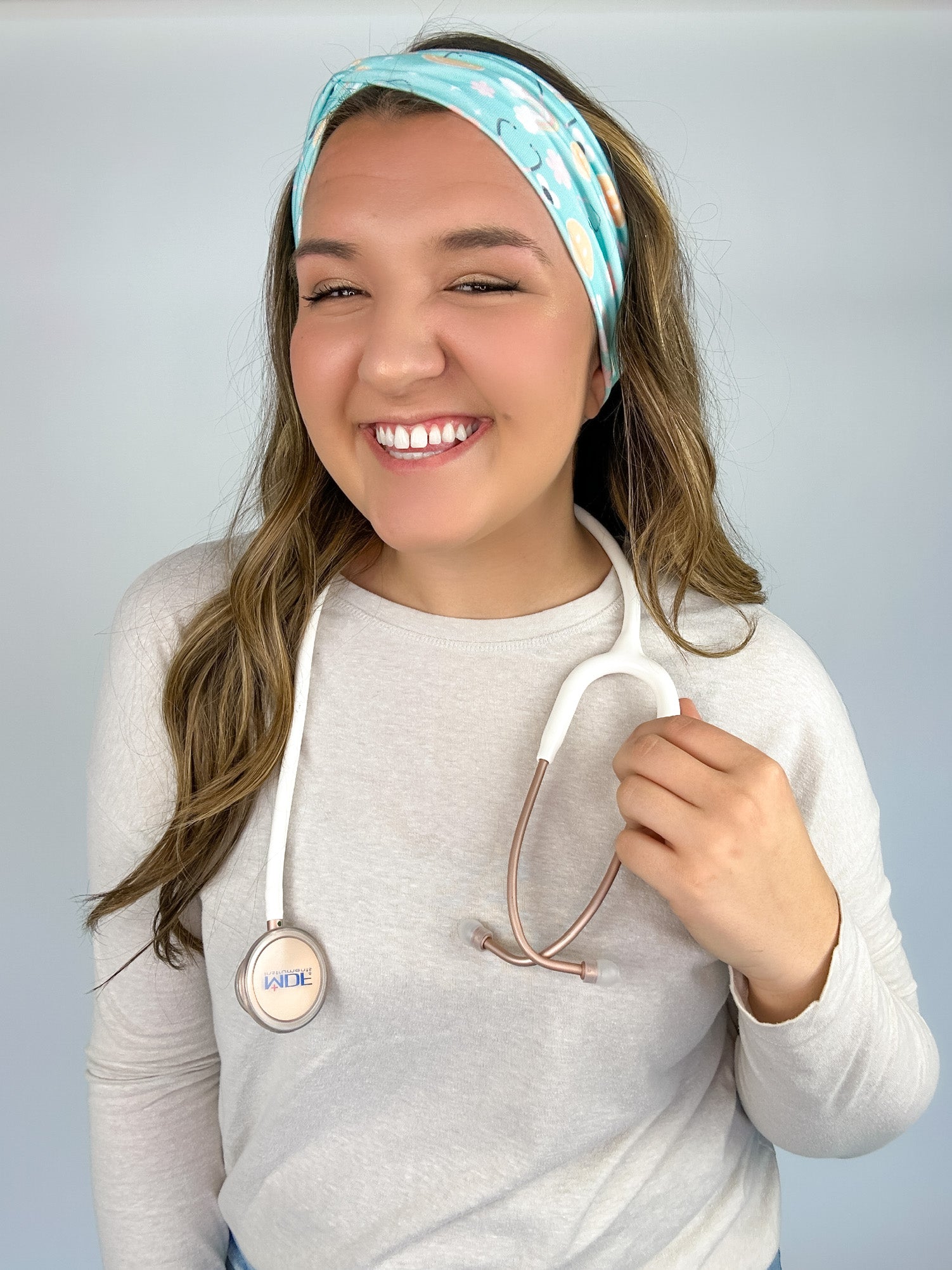 Adult women's blue-teal headband with yellow smiley faces, pink daisies, muted rainbows, and pink stethoscopes with a heart on the end