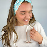 Adult women's blue-teal headband with yellow smiley faces, pink daisies, muted rainbows, and pink stethoscopes with a heart on the end