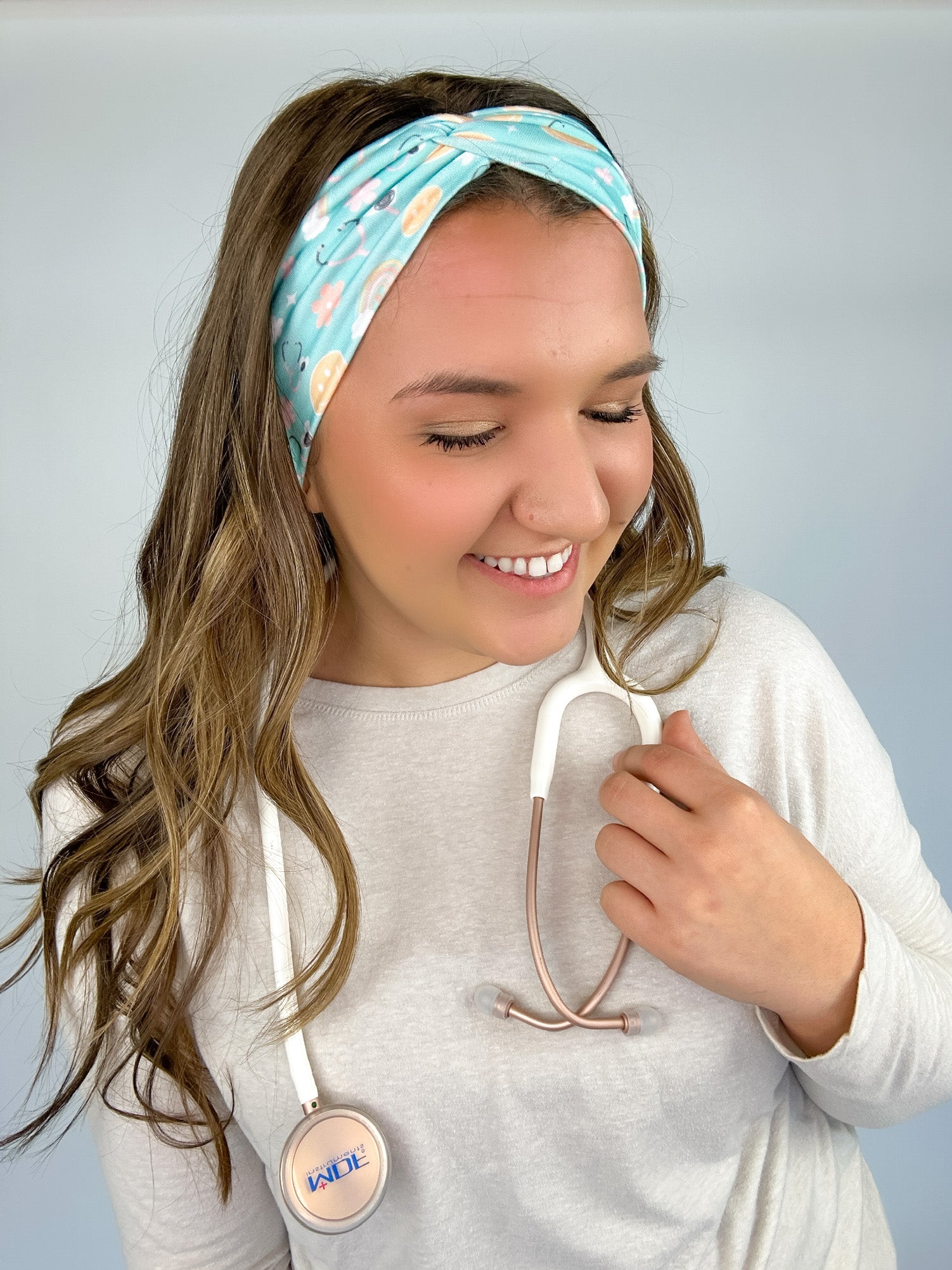 Adult women's blue-teal headband with yellow smiley faces, pink daisies, muted rainbows, and pink stethoscopes with a heart on the end