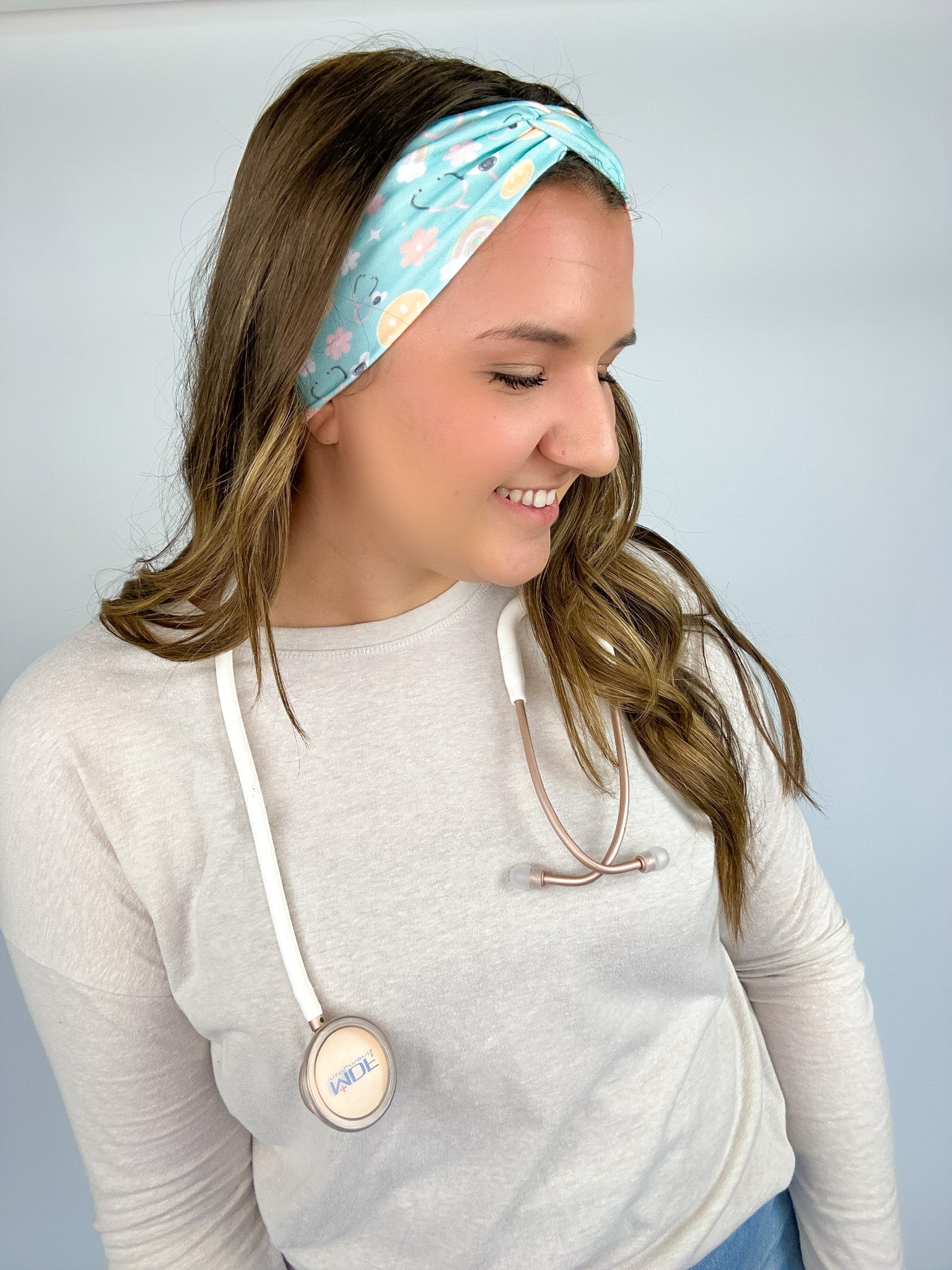 Adult women's blue-teal headband with yellow smiley faces, pink daisies, muted rainbows, and pink stethoscopes with a heart on the end