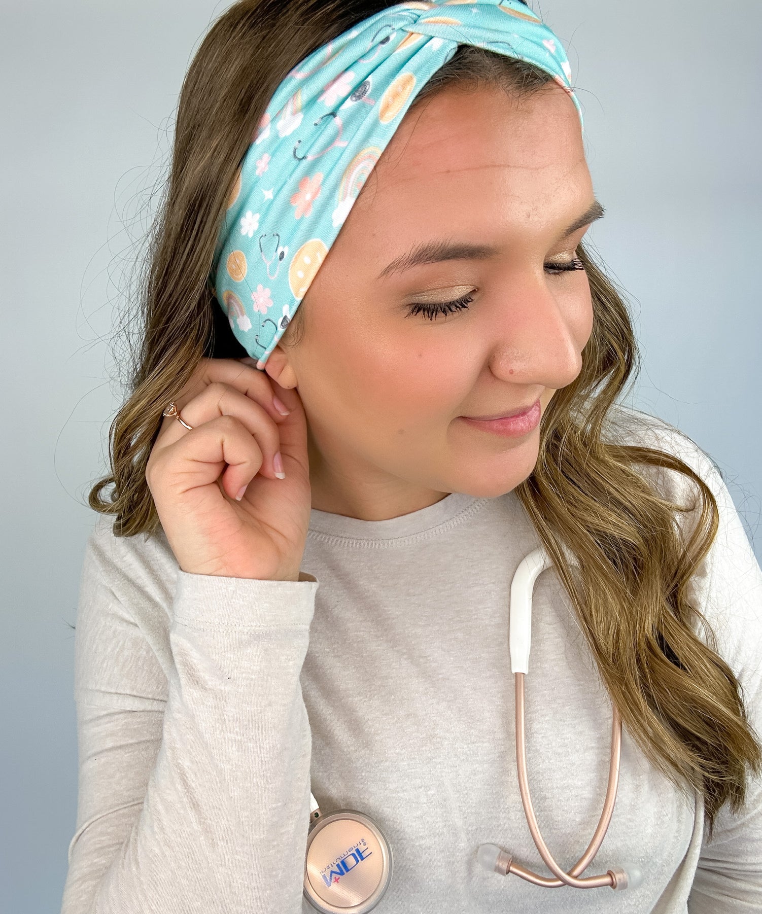 Adult women's blue-teal headband with yellow smiley faces, pink daisies, muted rainbows, and pink stethoscopes with a heart on the end