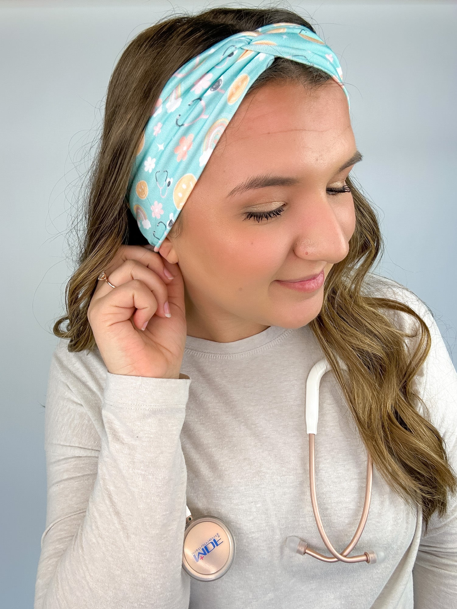 Adult women's blue-teal headband with yellow smiley faces, pink daisies, muted rainbows, and pink stethoscopes with a heart on the end
