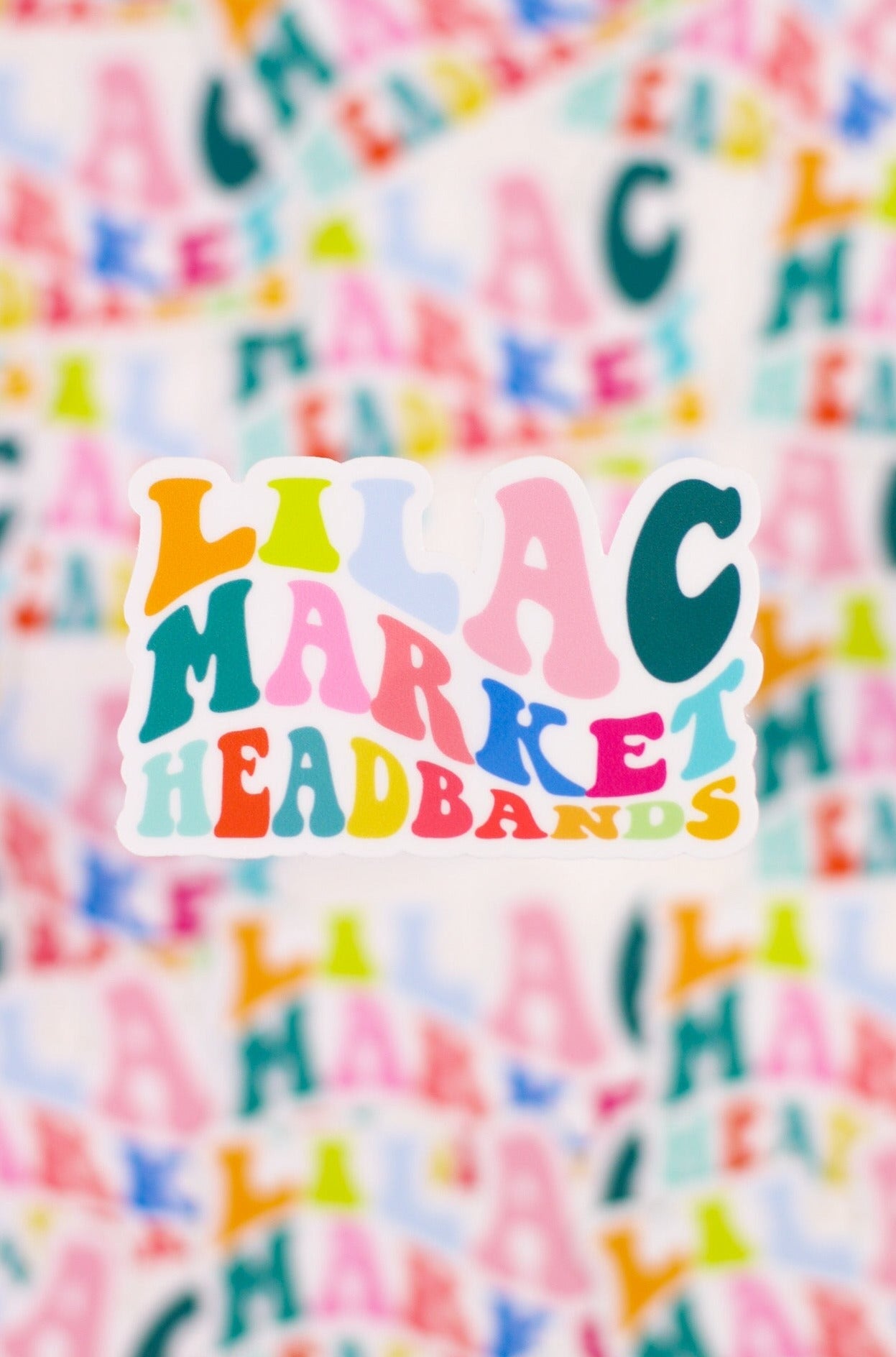 Decorative sticker with 70's style big and small font with the words "Lilac Market" with each letter a different color including orange, pink, teal, green, and blue.